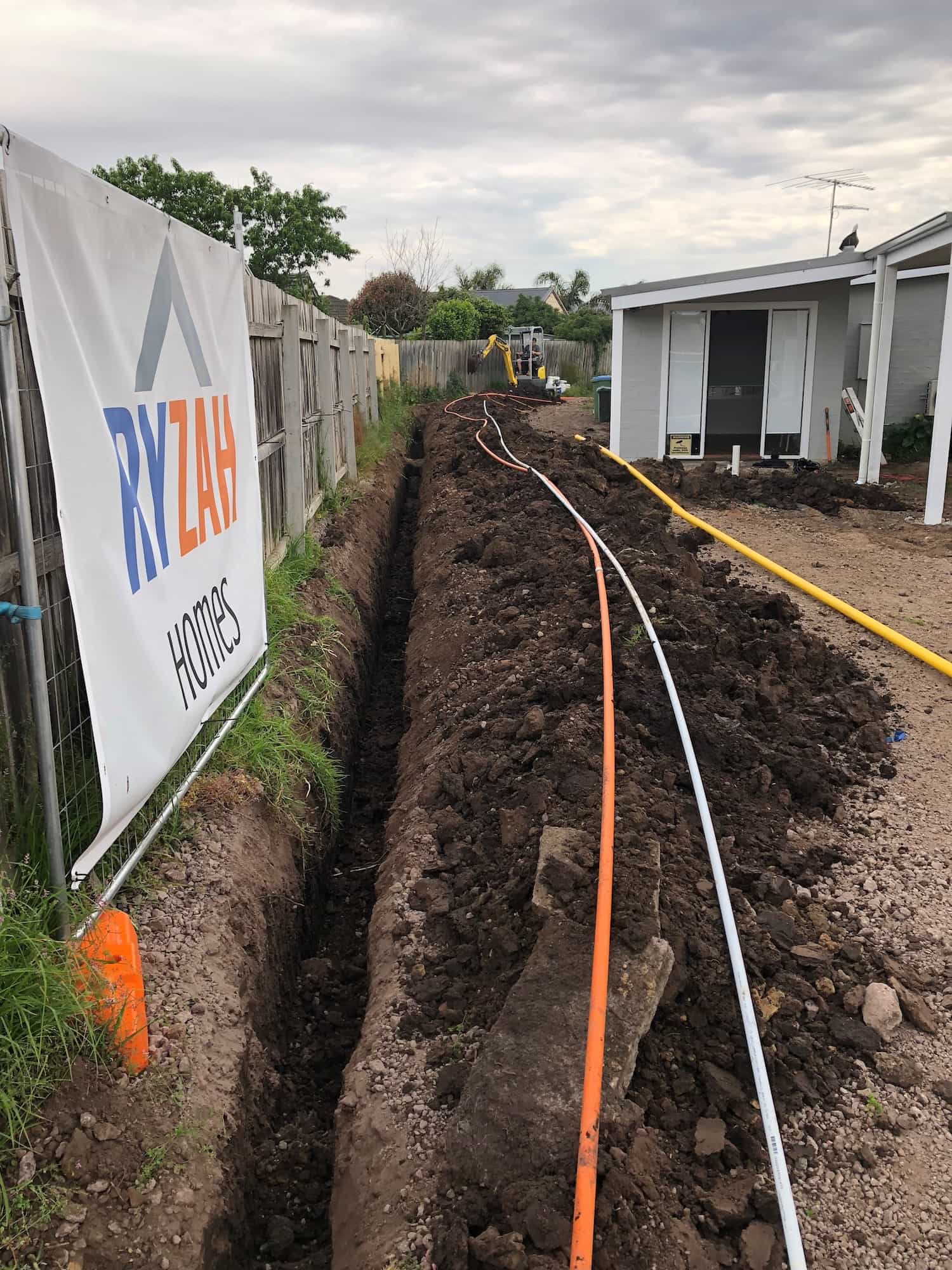 Excavated trench alongside a timber fence with orange, white and yellow service conduits laid ready for installation at a new residential build site, with a mini excavator and house in the background and a builder's hoarding sign in the foreground