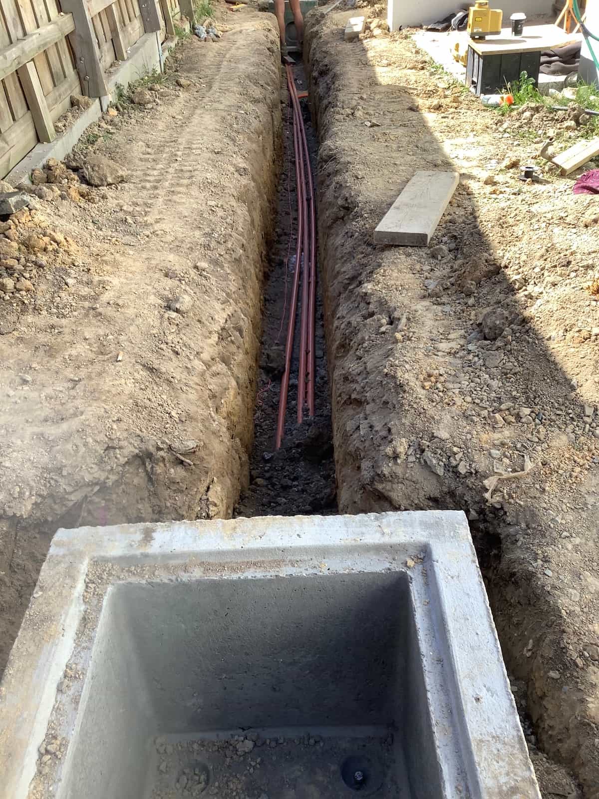 Close-up view of a narrow excavated trench containing multiple red electrical conduits leading into a precast concrete pit box in the foreground, at a residential construction site with timber fencing alongside